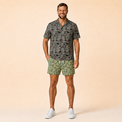 Man wearing Silpaz duck camo athletic shorts in sage green camo with a dark camp shirt, standing front view