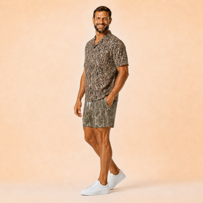 Man wearing Silpaz tree bark camo mesh shorts with a matching camp shirt, hand in pocket, full length view