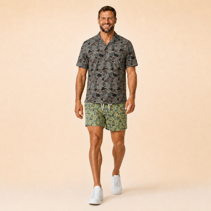 Man wearing Silpaz duck camo athletic shorts in sage green camo with a dark camp shirt, walking pose